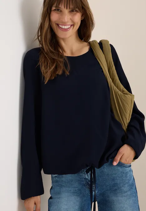 Frau lächelt, trägt dunkelblaues Oberteil, Jeans und eine olivgrüne Tasche.